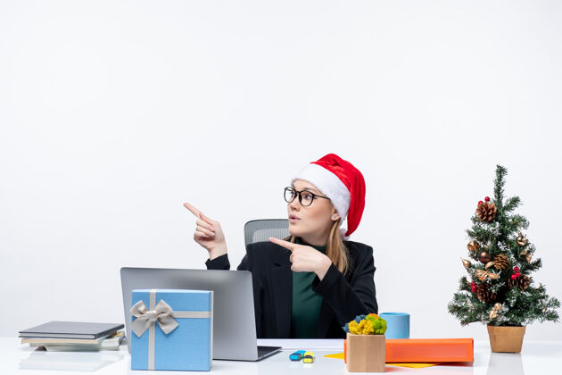 圣诞节新年气氛与年轻迷人的女人坐在一个圣诞树和礼物的桌子上 圣诞老人帽子集中在办公室的东西桌子帽子女人