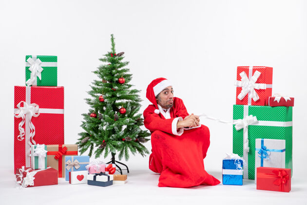 圣诞老人一个情绪激动的年轻人穿着圣诞老人的衣服 拿着礼物和装饰好的圣诞树坐在白色背景的地上装饰年轻人男人