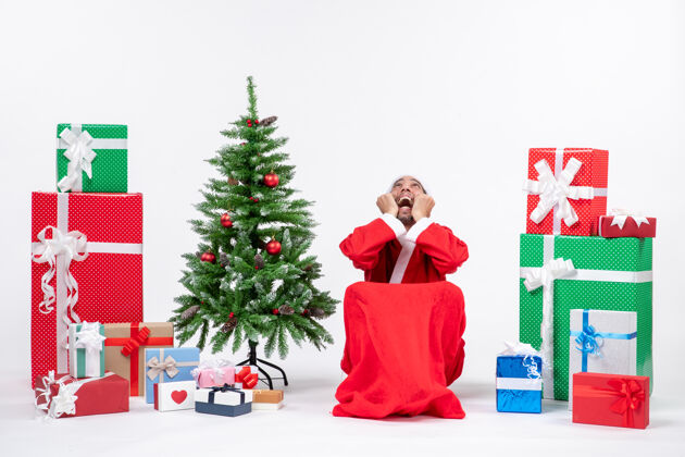 关心情绪激动的关心的年轻人打扮成圣诞老人与礼物和装饰圣诞树上的白色背景股票照片圣诞老人圣诞老人年轻人