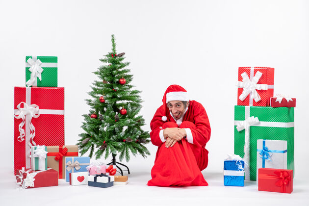 男人滑稽的年轻人打扮成圣诞老人 拿着礼物和装饰过的圣诞树坐在白色背景的地上年轻人圣诞老人圣诞树