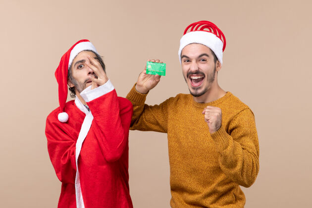 伙计们正面图两个快乐的家伙 一个穿着圣诞外套 手放在脸上 另一个拿着米色背景的卡片卡片外套圣诞老人