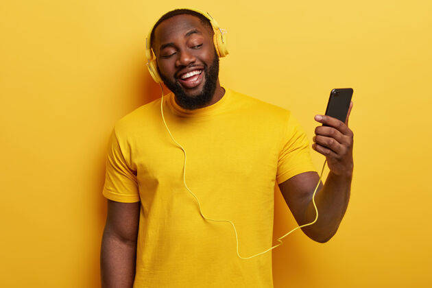 高兴快乐的非裔美国人喜欢唱歌 手持连接耳机的现代手机男性T恤胡须