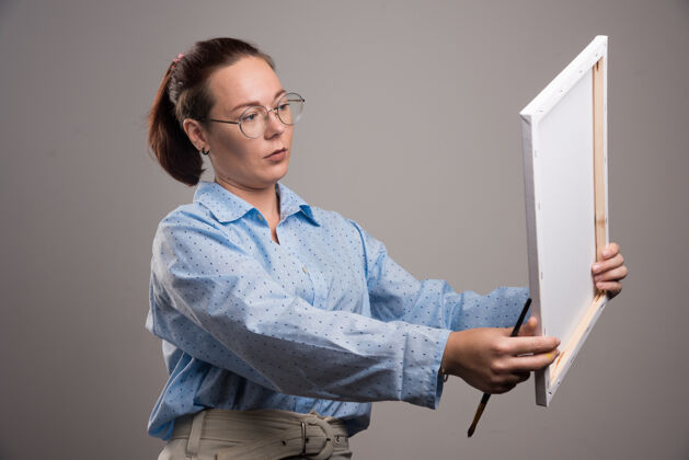 爱好戴眼镜的女人拿着帆布 在灰色背景上画笔高质量的照片持有女孩设备