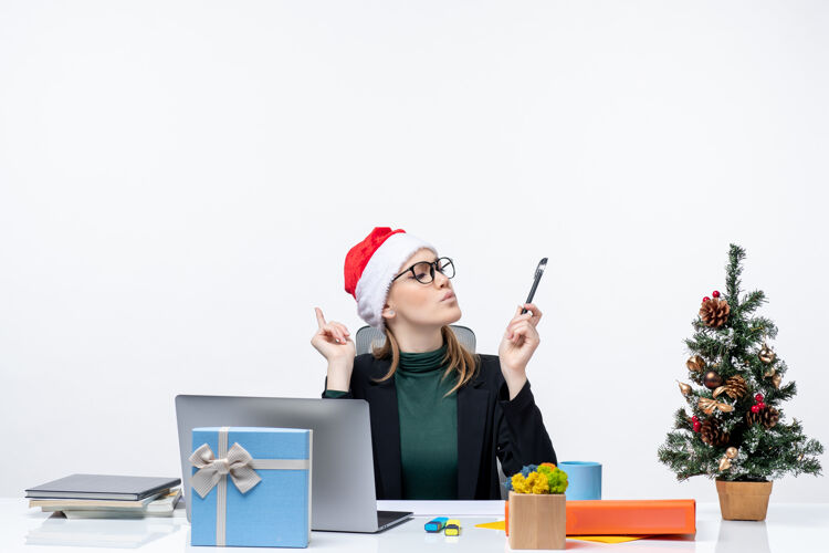女商人在白色背景的办公室里 一个戴着圣诞老人帽子的金发女人坐在一张桌子旁 桌子上放着圣诞树和礼物帽子圣诞节金发女郎
