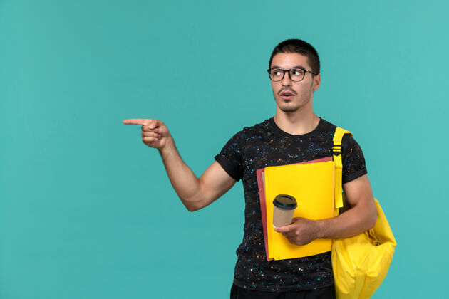 大学身穿深色t恤的男生正面图黄色背包 蓝色墙上放着不同的文件和咖啡背包男学生档案