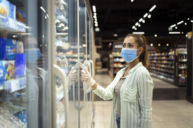 冰箱戴着防护口罩和手套的妇女在covid-19大流行或电晕病毒期间打开超市的冰箱食品杂货店持有病毒