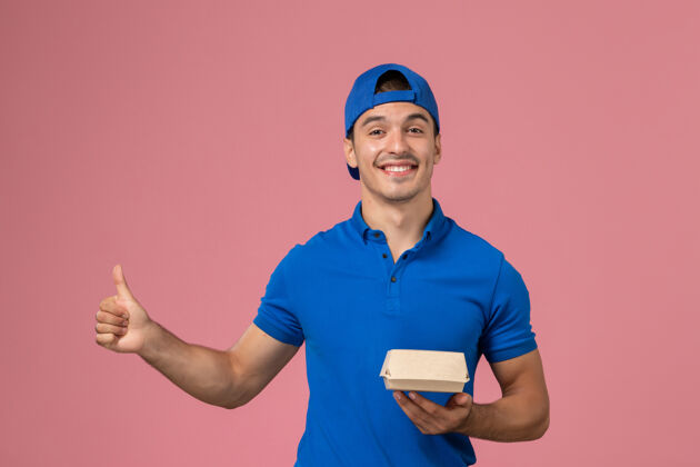 年轻人正面图：身穿蓝色制服披肩的年轻男性快递员手持粉色墙上的快递食品包男人粉色员工