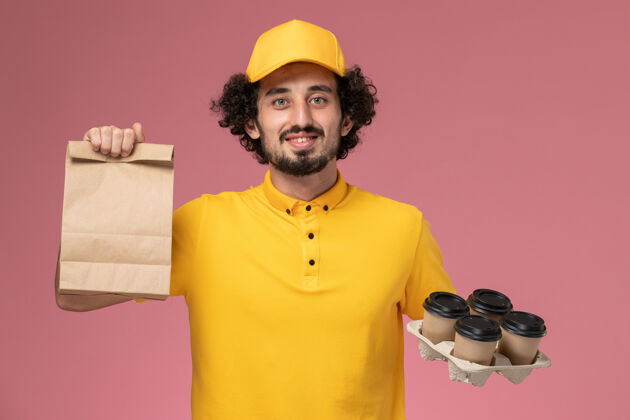 手持正面图：身穿黄色制服的男性快递员手持棕色的咖啡杯和食品包装放在粉红色的墙上正面男快递员黄色