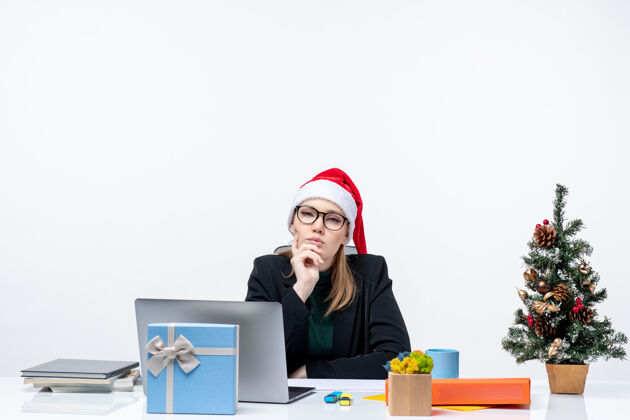 笔记本电脑新年的气氛与年轻迷人的女子在深深的思想与圣诞老人的帽子坐在一张桌子上 圣诞树和一个礼物在它的办公室桌子迷人圣诞节