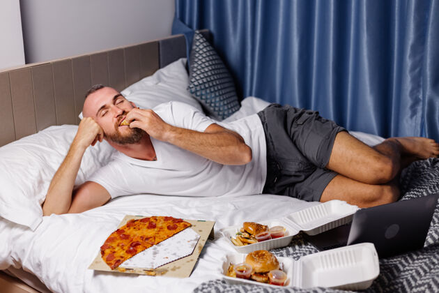 快餐快乐的白种人在家里卧室里吃快餐在床上男人在网上叫外卖吃比萨饼和汉堡在舒适的房间肖像小吃人