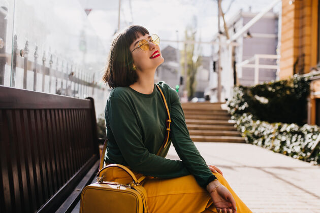 休息穿着新潮绿色毛衣的浪漫白种女孩在街上摆姿势幸福的深色女模特坐在长椅上的户外镜头休闲街道公园