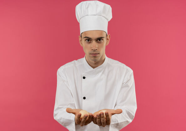年轻年轻的男厨师穿着厨师制服拿着鸡蛋站在隔离的粉红色墙上拿着制服厨师