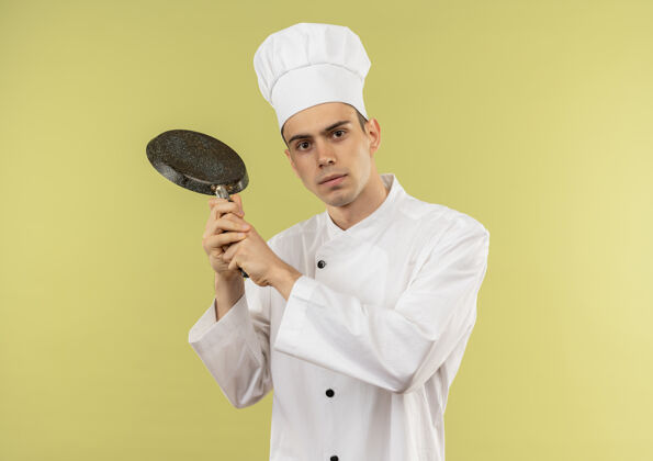 年轻年轻的男厨师穿着厨师制服 肩上扛着煎锅 站在隔离的绿色墙壁上 有复印空间肩膀看着平底锅