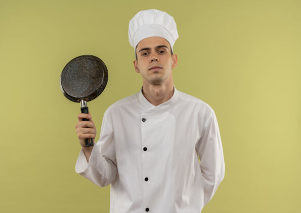 穿着年轻的男厨师穿着厨师制服 拿着煎锅站在隔离的绿色墙壁上 有复印空间拿着看着平底锅