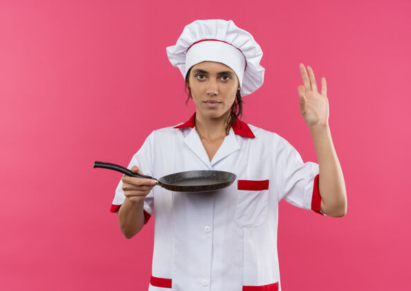 煎年轻的女厨师穿着厨师制服 拿着煎锅 在隔离的粉色墙壁上展示okey手势 还有复印空间拿着女好的