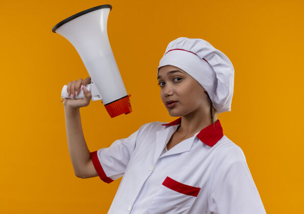 厨师年轻的女厨师穿着厨师制服拿着喇叭站在隔离的橙色墙上拿着年轻看着