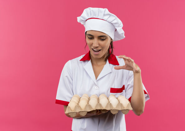 抱快乐的年轻女厨师穿着厨师制服 拿着和看一批鸡蛋在孤立的粉红色墙上复制空间鸡蛋厨师年轻
