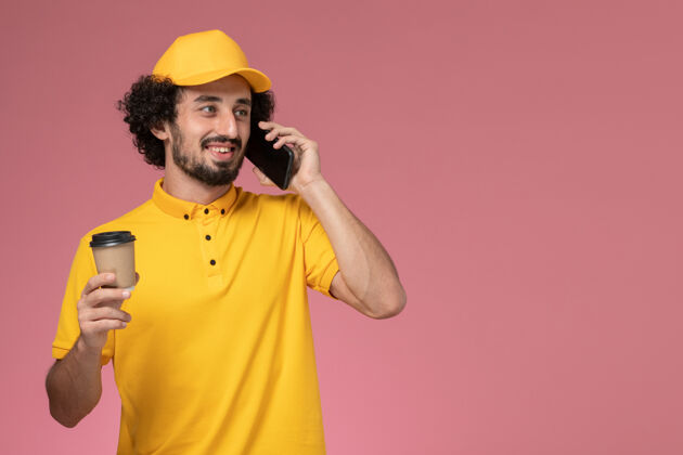杯子正面图身穿黄色制服的男信使和披风拿着送货咖啡杯在粉色墙上打电话手持谈话制服