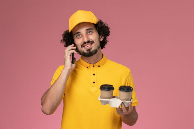 棕色正面图穿着黄色制服和披风的男信使拿着棕色的咖啡杯 在粉红色的墙上打电话咖啡服务工作