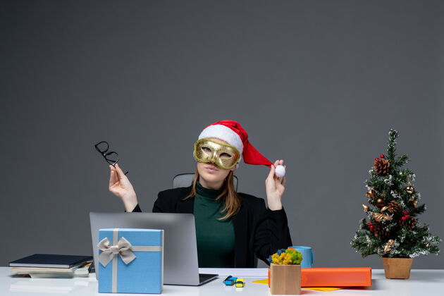 帽子圣诞节的心情与年轻女子玩圣诞老人戴着眼镜 戴着面具坐在一张桌子在黑暗的背景下的帽子办公室坐着笔记本电脑