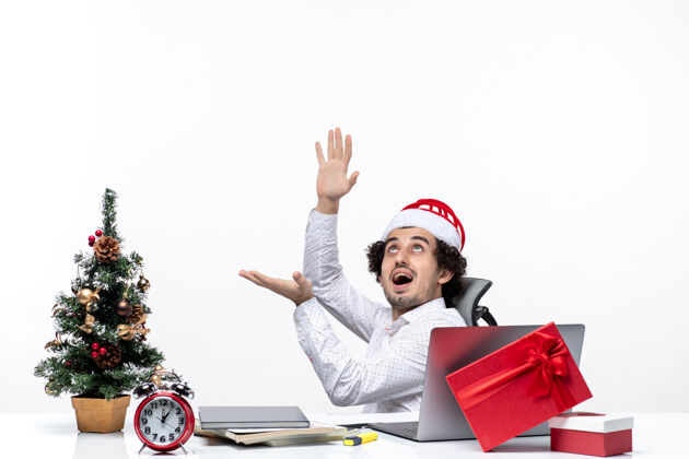 帽子带着微笑的新年心情积极的年轻商人戴着圣诞老人的帽子坐在办公室里 独自一人在白色背景下工作圣诞老人商人独自