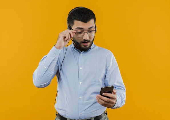 眼镜一个留着胡子的年轻人 穿着蓝色衬衫 戴着耳机 拿着麦克风 看着他的智能手机屏幕 摘下眼镜站着橙色放