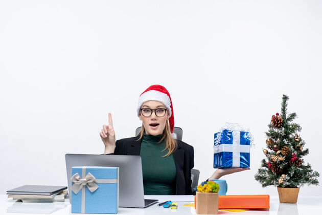 年轻女人带着圣诞老人帽子戴着眼镜的年轻女子坐在桌旁 在白色背景上指着上面的圣诞礼物 充满节日气氛笔记本坐着指着