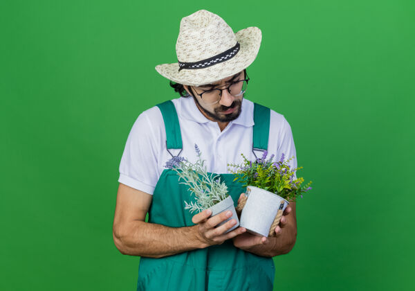 年轻年轻的留着胡子的园丁 穿着连体衣 戴着帽子 手里拿着盆栽植物 严肃地看着它们盆栽胡须站着