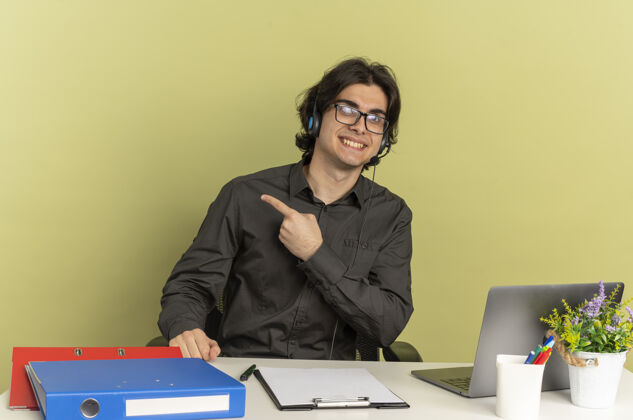 复制戴着耳机 戴着眼镜 面带微笑的年轻上班族坐在办公桌旁 办公工具放在旁边的笔记本电脑上工人微笑坐