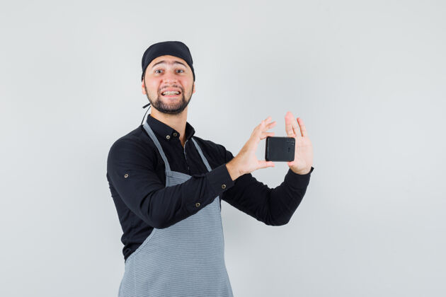 站男厨师穿着衬衫 围裙在手机上拍照 看上去很愉快正面图服用电话美食