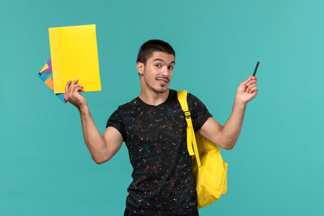 大学身穿深色t恤黄色背包的男学生正面图 浅蓝色墙上有文件和抄写本学院微笑人