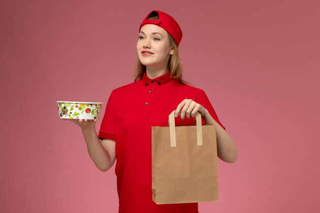快乐正面图：身穿红色制服 披风的年轻女快递员手持快递食品包和粉红色墙上的碗食物送货微笑