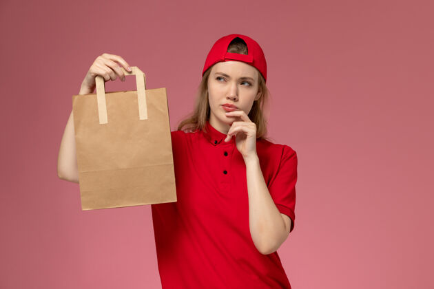 人正面图：身穿红色制服和披风的年轻女快递员手持快递食品包 在粉红色的墙上思考制服红色食物