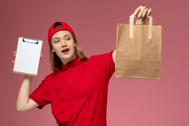 快乐正面图：身穿红色制服的年轻女快递员拿着快递食品包和笔记本放在浅粉色的墙上微笑女性制服
