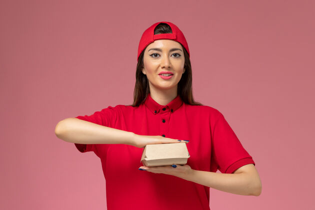 手正面图：身穿红色制服和斗篷的年轻女快递员 手上拿着一个小小的快递食品包 放在粉红色的墙上制服快人