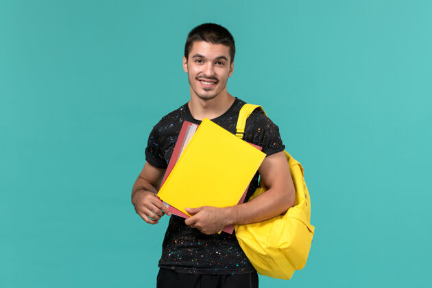 学生身穿深色t恤黄色背包的男学生正面图 浅蓝色墙上放着不同的文件学校成人人