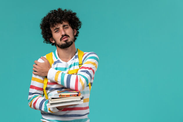 男学生身穿条纹衬衫 背着黄色背包 在蓝色墙上拿着书的男学生的正视图成人学校男