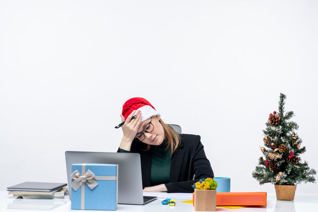 疲惫办公室里 一个疲惫过度的金发女人 戴着圣诞老人的帽子 坐在一张桌子旁 桌子上放着一棵圣诞树和一份礼物 背景是白色的女人帽子电脑