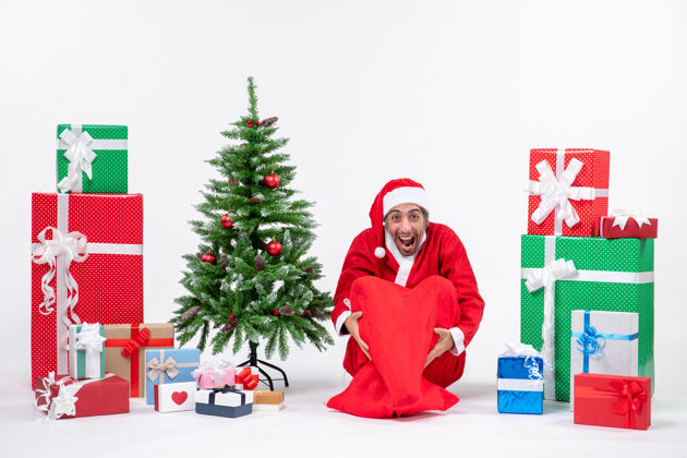 礼物快乐的年轻人装扮成圣诞老人 拿着礼物和装饰好的圣诞树坐在白色背景的地上礼物微笑人