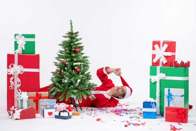 冬青节日气氛与年轻的惊喜圣诞老人躺在圣诞树后靠近礼物白色背景圣诞老人说谎礼物