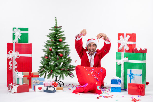 圣诞老人喜庆的节日气氛与有趣积极快乐的圣诞老人坐在地上 玩圣诞装饰品附近的礼物和装饰圣诞树上的白色背景人装饰圣诞老人