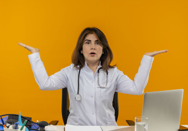 中年困惑的中年女医生穿着医用长袍 手持听诊器 坐在办公桌旁 拿着医疗工具在笔记本电脑上工作 双手放在隔离的橙色墙壁上 留有复印空间女性穿着手