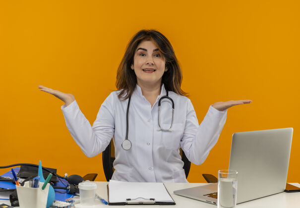 坐着穿着医用长袍 手持听诊器的中年女医生坐在办公桌旁 拿着医疗工具在笔记本电脑上工作 双手放在隔离的橙色墙壁上 留有复印空间笔记本电脑工具医疗