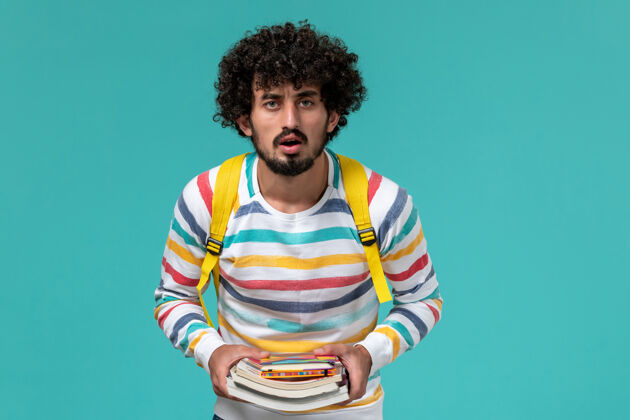 画像穿着条纹衬衫 背着黄色背包 拿着书的男学生在蓝色墙上的正视图条纹学生男