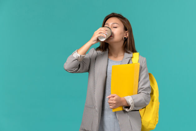饮料身穿灰色夹克 背着黄色背包 拿着文件 在淡蓝色墙上喝咖啡的女学生的正视图大学Female文件