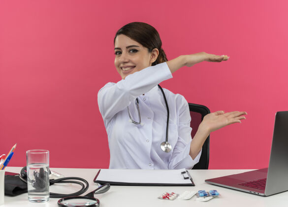 坐着微笑的年轻女医生穿着医用长袍带听诊器坐在办公桌前用医疗工具在电脑上工作显示大小和复印空间工作展示大小