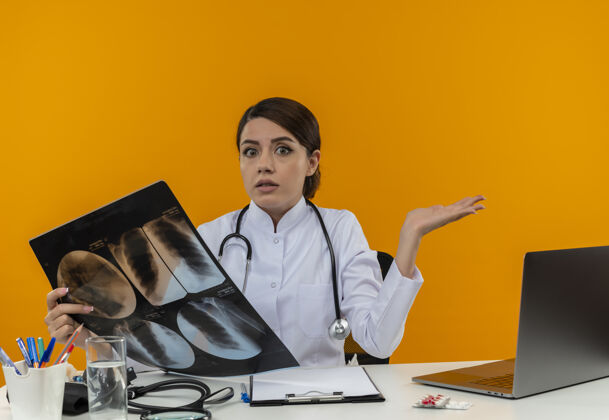拿着困惑的年轻女医生穿着医用长袍 手持听诊器 坐在办公桌旁 拿着医疗工具 拿着x光片在黄色隔离墙上工作医疗困惑穿着