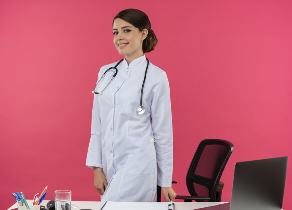办公桌面带微笑的年轻女医生穿着医用长袍 手持听诊器 站在办公桌后面 用带复印空间的医疗工具在电脑上工作电脑工具女