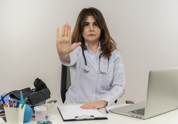女性严格的中年女医生 穿着医用长袍 带听诊器 坐在办公桌旁 用笔记本电脑和医疗工具 在隔离的白色背景上显示停止手势 并留有复印空间中年医生长袍