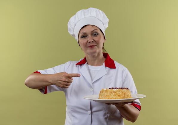 高兴一位身着厨师制服的中年女厨师手拿蛋糕放在盘子里 手指指向一边 站在隔离的绿色墙壁上蛋糕厨师点